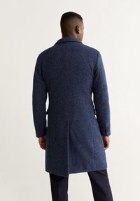 Cappotto in lana blu navy con texture a spina di pesce, maniche lunghe e colletto a revers; presenta due tasche laterali e bottoni ai polsini.