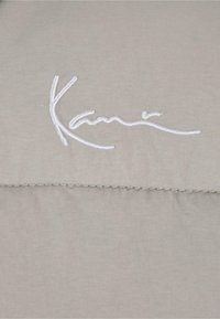 Svijetlo siva tkanina s glatkom teksturom ima bijeli izvezeni tekst "Kari" i ušivan detalj šava.