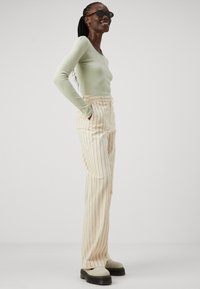 Light green ribbed long-sleeve top paired with cream striped wide-leg pants and chunky cream shoes. Model poses with hands in pockets.