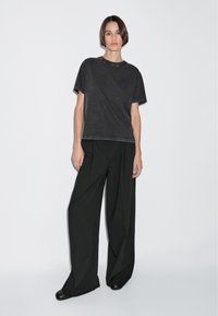 Maglietta nera oversize con collo a giro e bordi grezzi, abbinata a pantaloni neri a gamba larga con pieghe e una texture liscia.