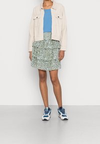 Svetle béžová kordurová bunda se dvěma předními kapsami, kombinovaná s modrým tílkem a sukní s vrstveným zeleným květinovým vzorem. Bílé tenisky.