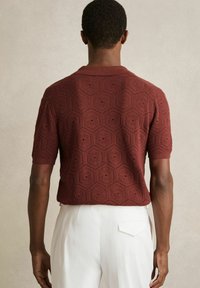 Polo din tricot de culoare maro cu mânecă scurtă, cu un model hexagonal și o suprafață texturată; are un guler tradițional. Stilizat cu pantaloni albi.