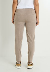 Pantalons beige en chevron avec une coupe ajustée, dotés de deux poches arrière et d'une texture lisse, associés à des baskets beiges et métalliques.