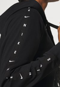 Personne portant un sweat à capuche noir Nike avec un logo blanc et des inscriptions le long de l'épaule et de la manche, tenant le cordon de la capuche.