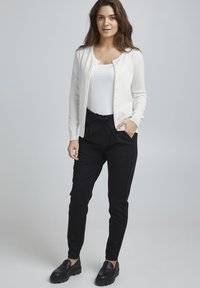 Cardigan bianco lavorato a maglia con scollo rotondo, chiusure a bottoni, indossato sopra una canotta bianca e pantaloni neri con coulisse. Scarpe nere senza lacci.
