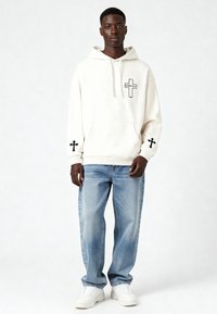 Felpa crema con tasca frontale e design a croce nera su maniche e petto, abbinata a jeans blu chiaro e sneakers bianche.