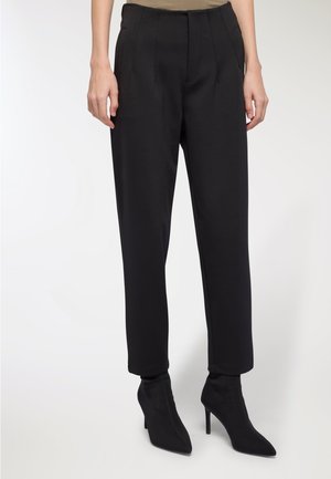 Pantaloni neri su misura con vita alta, caratterizzati da pieghe e tasche laterali, abbinati a stivaletti neri con tacco.