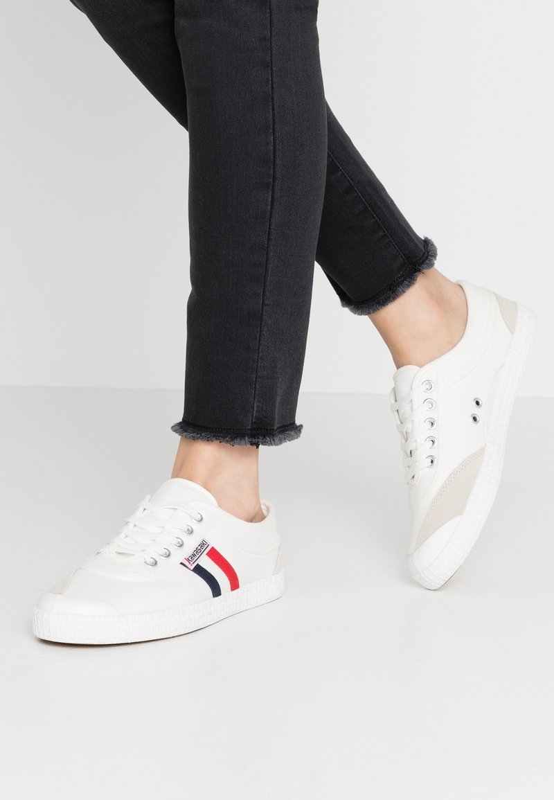 White canvas sneakers featuring a rubber sole, red and navy stripes, and gray suede accents. Laces and eyelets visible. Casual design.