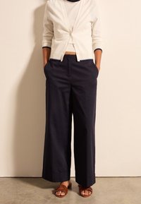 Osoba ubrana w biały krótką kardigan, szerokie granatowe spodnie z rękami w kieszeniach oraz brązowe plecione sandały stoi na betonowej podłodze.