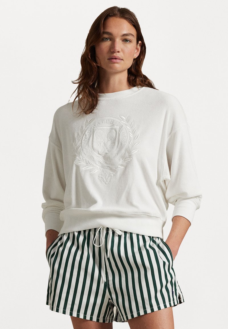 Hvit sweatshirt med preget logo og emblem på forsiden, kombinert med grønne og hvite stripete shorts, som har sidelommer og snøring.