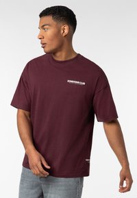 Bordeauxrotes T-Shirt mit kurzen Ärmeln aus Baumwolle, mit Rundhalsausschnitt, einem Logo-Text auf der Vorderseite und einer lässigen Passform. Kombiniert mit hellgrauen Jeans.