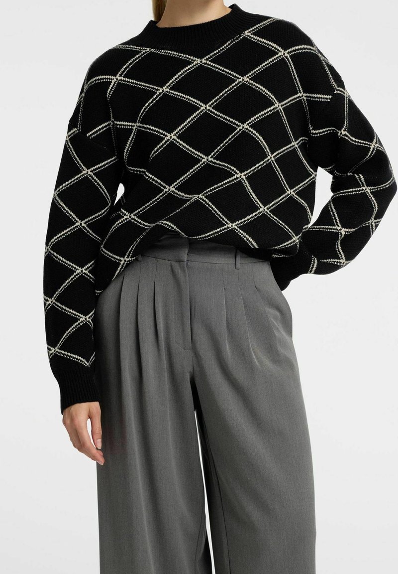 Svart stickad tröja med vitt diamantmönster, ribbat halsband och långa ärmar. Matchad med grå veckade byxor.