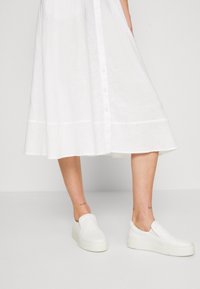 Vestido blanco abotonado con falda fluida, combinado con zapatillas blancas sin cordones con suela texturizada y collar acolchado.