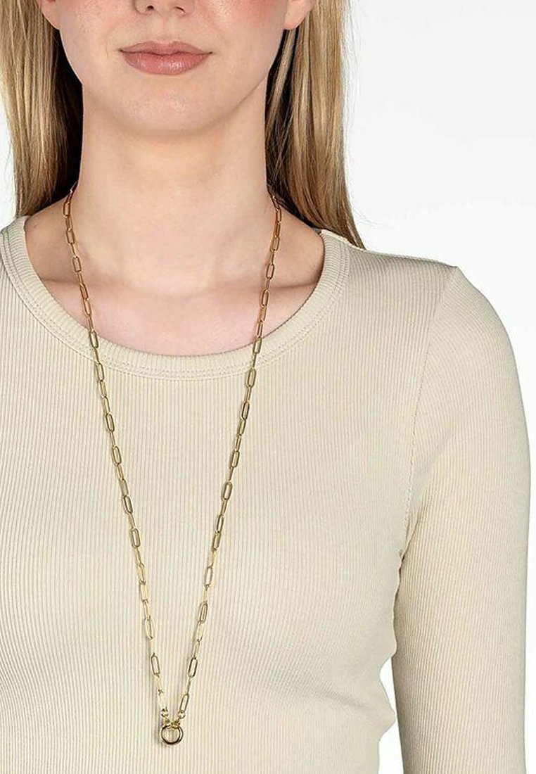 Guldkæde med forlængede led og en rund vedhæng. Båret over en lysebeige ribbet top, der viser et enkelt design.