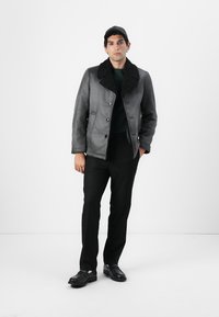Giacca shearling grigia con colletto nero testurizzato, dotata di bottoni, indossata sopra un maglione scuro e abbinata a pantaloni neri e scarpe.