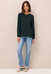 Blusa verde scuro a maniche lunghe con scollatura arricciata e dettagli di nodo, abbinata a jeans a gamba dritta blu chiaro e sneakers beige con accenti verdi.