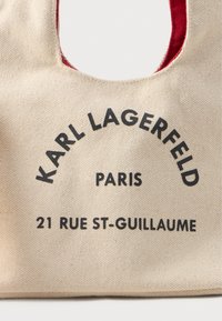 Geantă de toaleta din pânză bej cu interior roșu, text negru "KARL LAGERFELD PARIS 21 RUE ST-GUILLAUME" imprimat pe față.