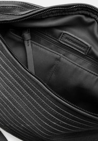 Innenansicht einer schwarzen Leder Tasche, die eine strukturierte Oberfläche mit diagonaler Naht, einer Reißverschlusstasche und einem Markenlabel zeigt.