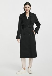 Czarny płaszcz trench z podwójnymi guzikami, regulowanym paskiem i naramiennikami, zestawiony z jasnymi spodniami i białymi trampkami.