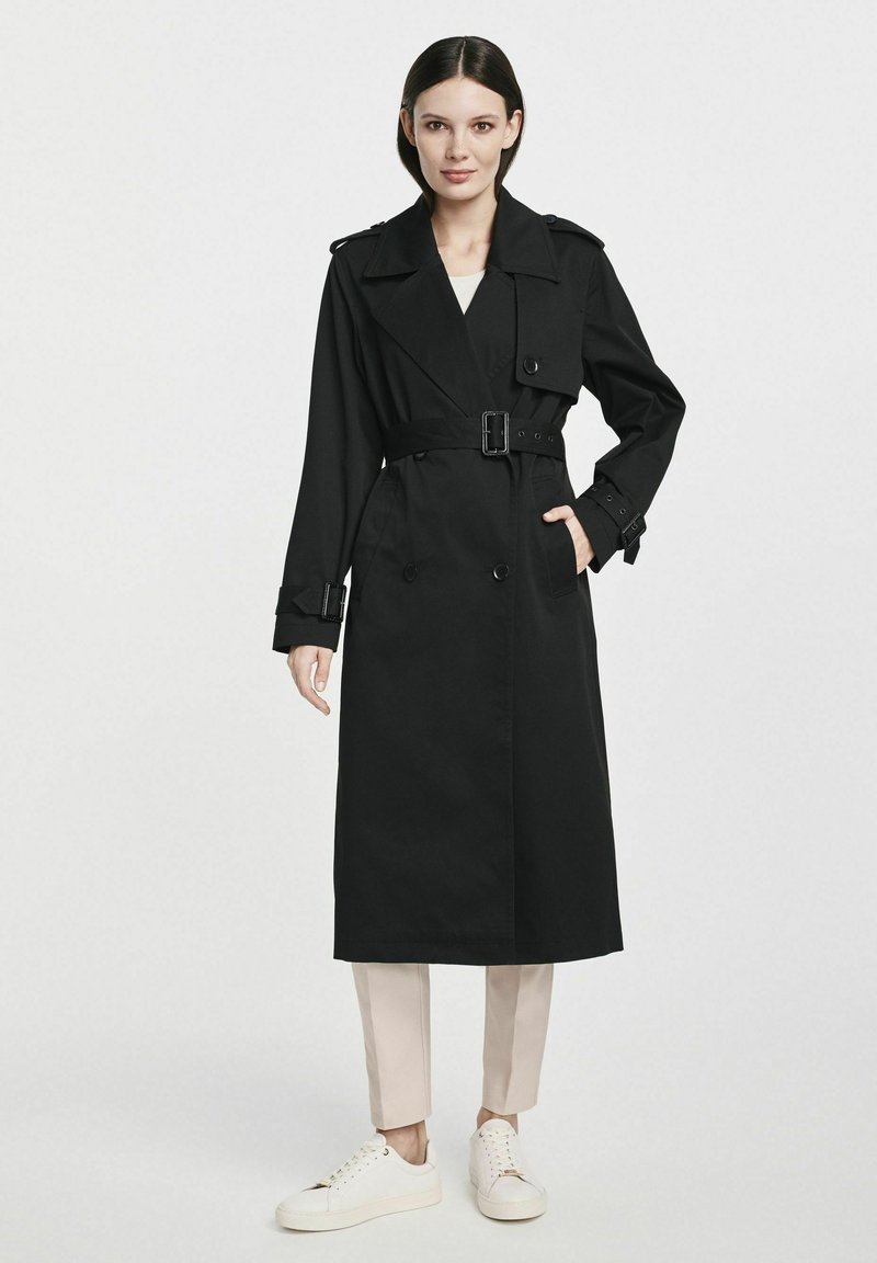 Czarny płaszcz trench z podwójnymi guzikami, regulowanym paskiem i naramiennikami, zestawiony z jasnymi spodniami i białymi trampkami.