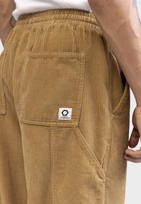 Tan corduroy pants with a textured ribbed pattern, featuring side pockets and a small logo label on the back pocket. Elastic waistband visible.