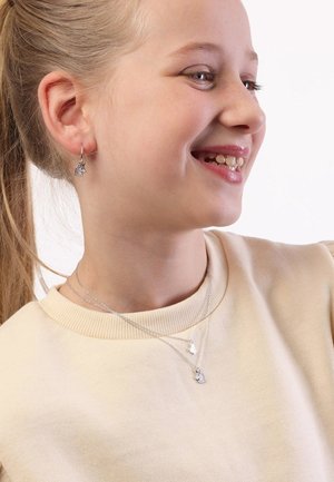 Zilveren eenhoorn oorbellen en gelaagde zilveren kettingen gedragen door een meisje in een beige sweatshirt met een ribbelachtige halslijn. Gladde texturen zichtbaar.