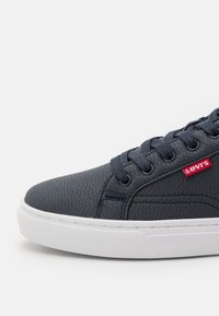 Sapatilha de couro texturizado azul escuro com sola de borracha branca. Apresenta atacadores redondos e um patch do logótipo vermelho da Levi's na lateral.