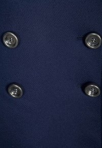 Tmavě modrý kabát na dvouřadé zapínání se stříbrnými knoflíky, měkkou vlněnou texturou a čistým designem s minimálními detaily na šití.