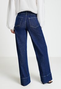 Pantaloni di jeans blu a gamba larga con finitura sbiadita, dotati di due tasche posteriori e orlo risvoltato, abbinati a una maglia bianca a maniche lunghe e ampia.