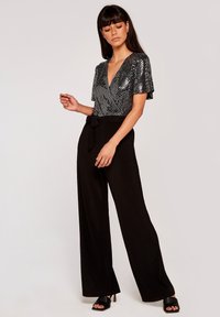 Bodysuit negro con lentejuelas y mangas cortas tipo volante, escote en V profundo, combinado con pantalones negros de talle alto y pierna ancha y sandalias de tacón negras.