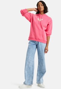 Sudadera rosa con bolsillo frontal y logo, combinada con vaqueros de pierna ancha en un tono azul claro. Zapatillas blancas completan el conjunto.