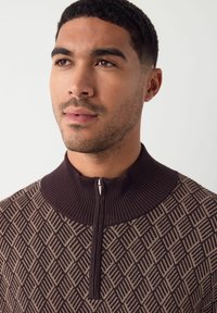 Braun gemusterter Pullover mit hohem Rippkragen und einem halben Reißverschluss, der ein Diamantmuster in kontrastierenden Beigetönen zeigt.