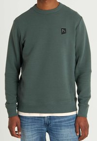Groene geribbelde sweatshirt met een ronde hals, voorzien van een klein zwart logo-embleem op de borst en lange mouwen. Gedragen over een wit shirt.