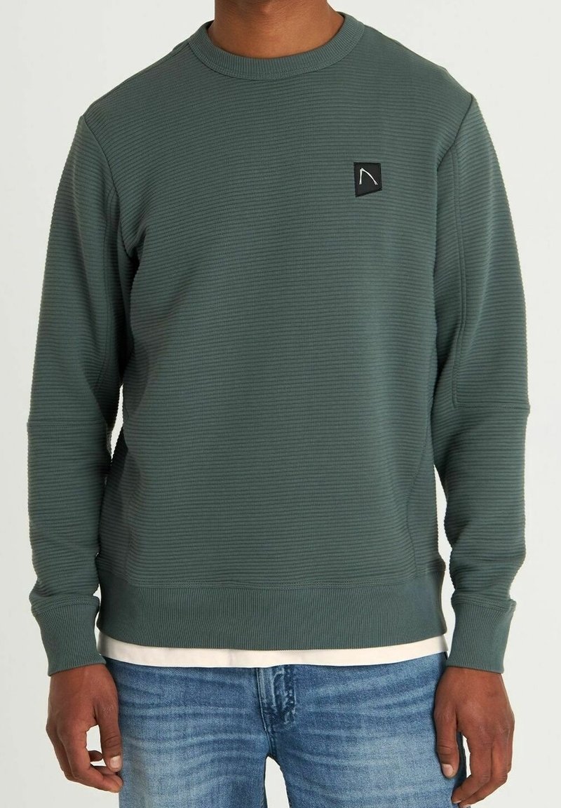 Groene geribbelde sweatshirt met een ronde hals, voorzien van een klein zwart logo-embleem op de borst en lange mouwen. Gedragen over een wit shirt.