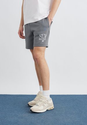 Person trägt graue New Balance-Shorts, weiße Socken und beige New Balance-Sneaker und steht auf einem blauen Teppich.