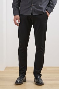 Pantalons noirs slim-fit, tissu lisse, design fuselé, avec poches latérales. Associés à des mocassins noirs et une chemise grise à boutons.