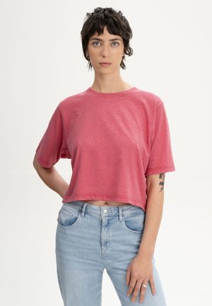 Jong persoon met kort, donker haar draagt een roze crop top en lichtblauwe jeans, staand tegen een effen witte achtergrond.