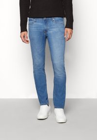 Man med slim-fit ljusblå jeans, vita sneakers och en mörk långärmad tröja, stående mot en enkel vit bakgrund.