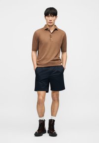Homme portant un polo tricoté à manches courtes marron, un short bleu marine, des chaussettes blanches et des bottines à lacets marron foncé, debout les mains dans les poches sur fond blanc.