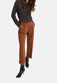 Pull noir à texture avec des manches longues, associé à un pantalon marron ajusté et des bottines noires en cuir verni à bout pointu.