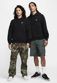 Μαύρες φούτερ με κουκούλα και μπροστινές τσέπες φορεμένες πάνω από καμουφλάζ παντελόνια cargo και πράσινα σορτς. Αθλητικά παπούτσια και λευκές κάλτσες μέχρι τον αστράγαλο ολοκληρώνουν τα σύνολα.