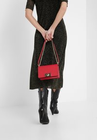 Rød rektangulær håndtaske med kæde-strop, har et metallisk spænde og holdes bag en person iført en glitrende sort kjole og højhælede støvler.