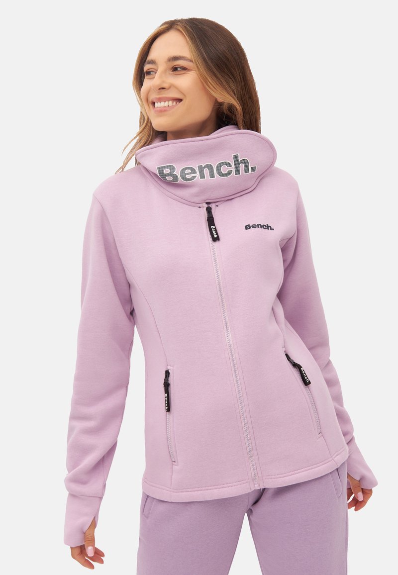 Light purple zip-up jacket with a high collar, featuring black logo accents and zippered pockets. Smooth, soft fabric with ribbed cuffs.