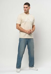 Béžové tričko s krátkym rukávom a veľkým bielym logom. Nosené s modrými džínsami s rovnými nohavicami a bielymi teniskami. Pozadie je svetlosivé.