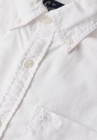 Camisa de algodón blanca con un diseño de botones, que presenta una textura suave, un logo bordado en el bolsillo del pecho y botones de plástico transparente.