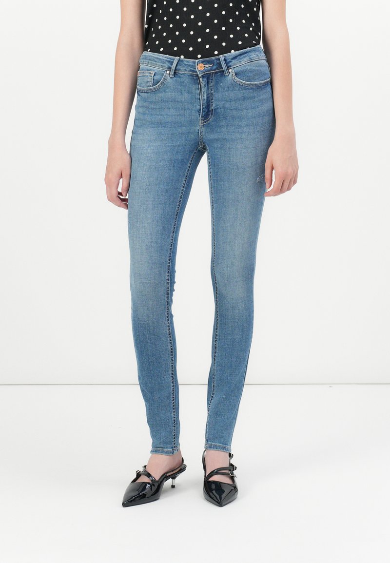 Slim-fit blue denim jeans with a five-pocket design, light wash, and subtle fading, paired with black polka dot top and black heeled shoes.