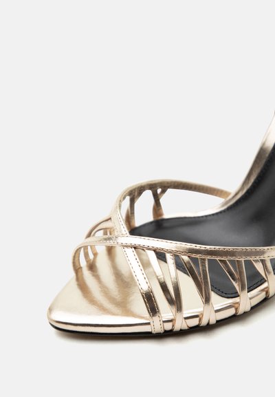 Χρυσό σανδάλι με ψηλό τακούνι και λουράκια, ανοιχτό στη μύτη, με πολλαπλά λεπτά λουράκια πάνω σε μαύρη σόλα, σε λευκό φόντο.