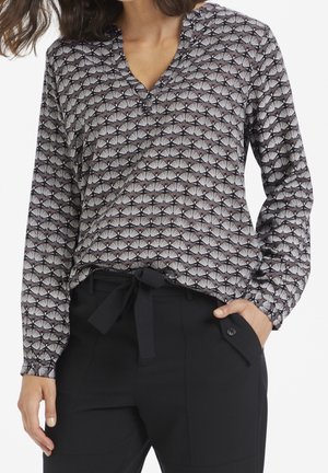 Vrouw draagt een blouse met lange mouwen, een patroon en een V-hals, gecombineerd met zwarte hoge taillebroek met een gestrikte ceintuur en een zak aan de zijkant met knopen.