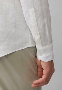 Weißes Leinenhemd mit knöpfbarem Manschette. Der Stoff ist leicht und strukturiert, mit einer geschwungenen Kantenmanschette und einem einzelnen Knopfverschluss.