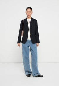 Marineblazer met gouden knopen, witte t-shirt met logo en lichtblauwe bredere jeans. Zwarte platte schoenen maken de outfit compleet.
